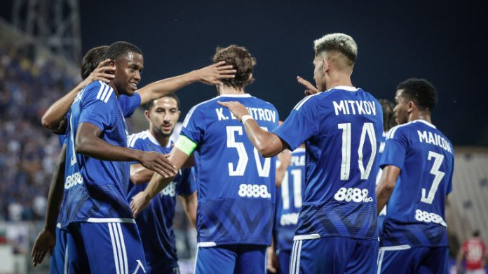 Soccer players in blue jerseys huddle and celebrate on the field at night.