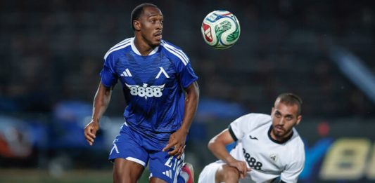 Левски поздрави Алдаир за рождения му ден Two soccer players on a field chase a ball in midair; blue jersey leads while a white jersey pursues.