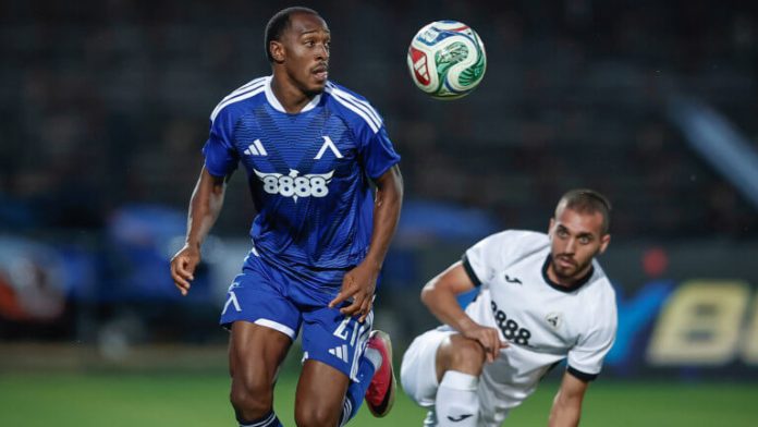 Two soccer players on a field chase a ball in midair; blue jersey leads while a white jersey pursues.