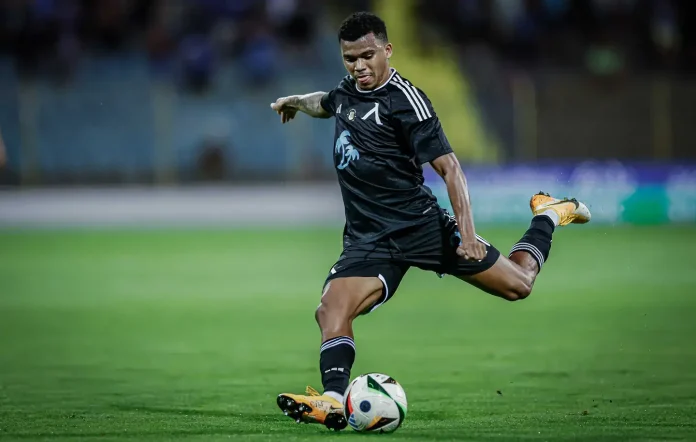 Soccer player in black kit kicking a white ball on a green field, right leg extended in a dynamic strike