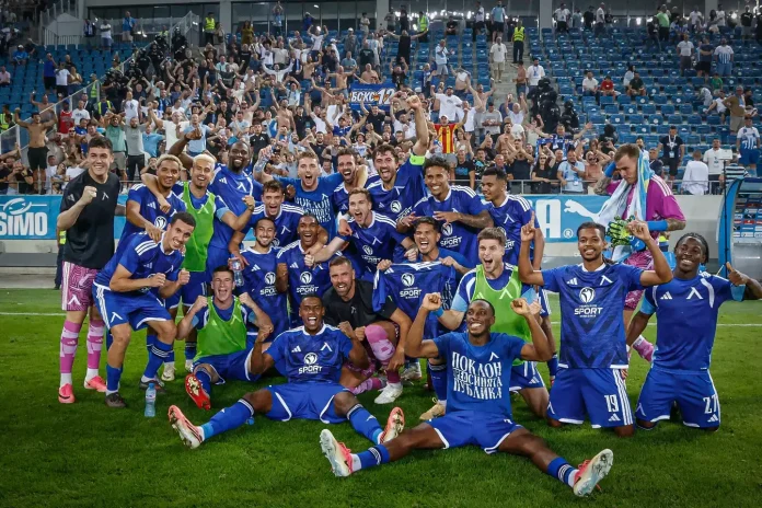 Soccer team in blue uniforms celebrates on the field with arms raised, as fans cheer from the stands behind them.] ,