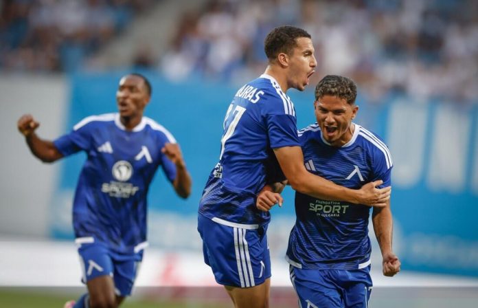 Three soccer players in blue uniforms celebrate on the field, embracing and cheering.