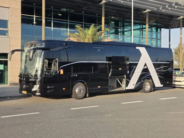 Black luxury tour bus with a large white A design on its side, parked in front of a glass-fronted building.