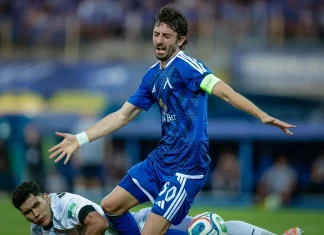 В Левски притеснени за Кристиан Димитров Male soccer player in blue kit with a captain's armband stretches his arms during a match, while a white-uniform opponent lies on the ground nearby.