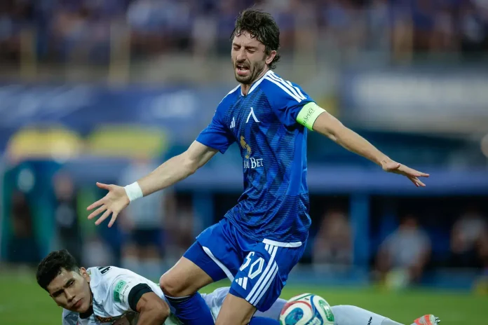 Male soccer player in blue kit with a captain's armband stretches his arms during a match, while a white-uniform opponent lies on the ground nearby.