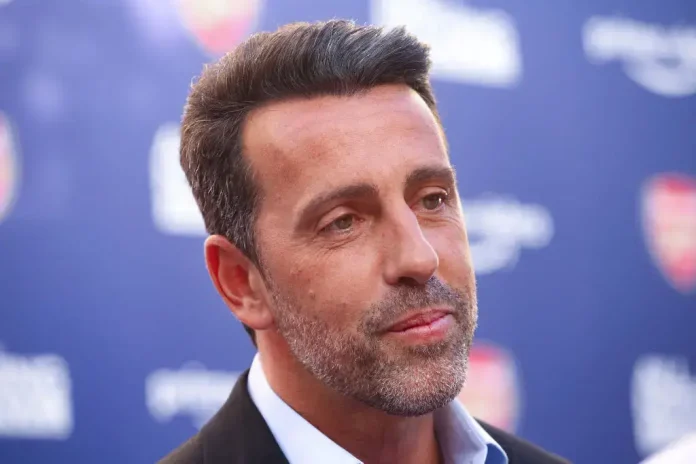Close-up of a man with salt-and-pepper stubble and dark hair in a suit, posing at a blue backdrop event photo backdrop behind him