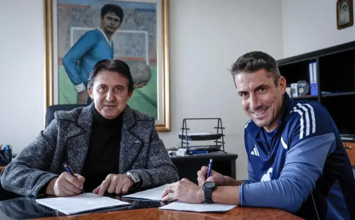 Two men sit at a desk signing documents; one in a tweed jacket, the other in a blue athletic top, smiling at the camera.