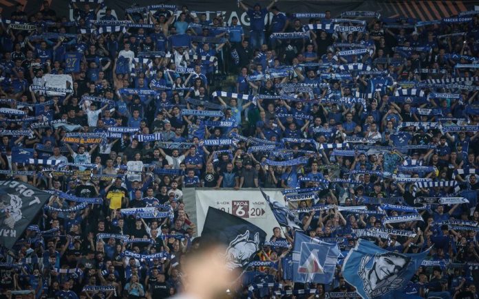 Large crowd of football fans in blue, waving scarves and banners in stadium stands in support of their team.