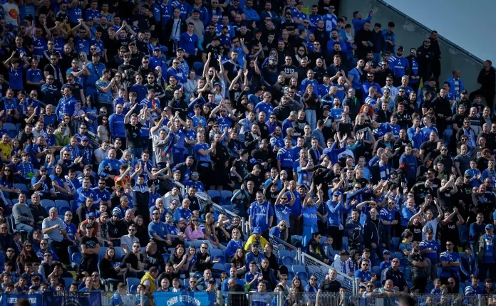 Large crowd of sports fans in blue, seated in a stadium, many clapping and cheering.