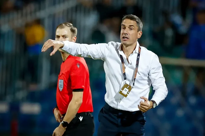 Soccer coach in a white shirt on the sideline, gesturing with his arm while a referee in red stands nearby during a match.