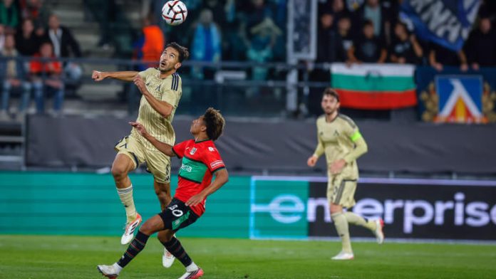 Two players jump to head the ball during a soccer match, one in beige and the other in red and black.