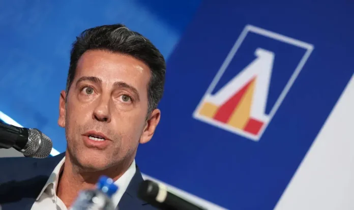 Man speaking into a microphone at a conference, with a blue backdrop and a geometric logo nearby