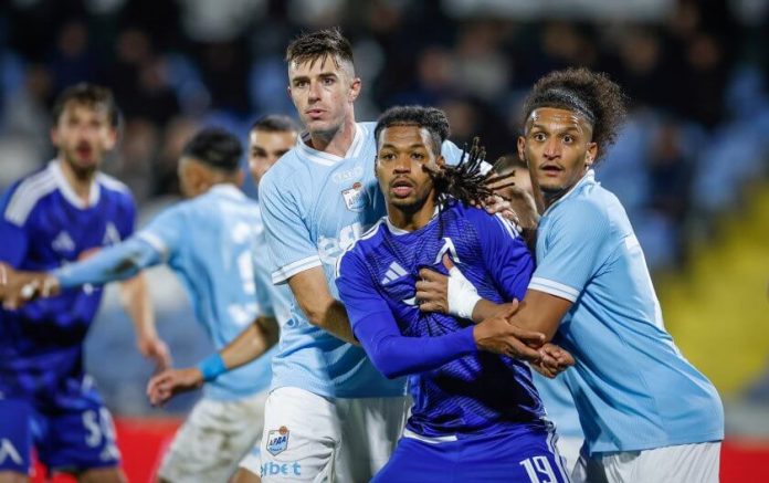 Three soccer players battle for the ball: two in light-blue jerseys brace a purple-clad player as they contest near the goal area.