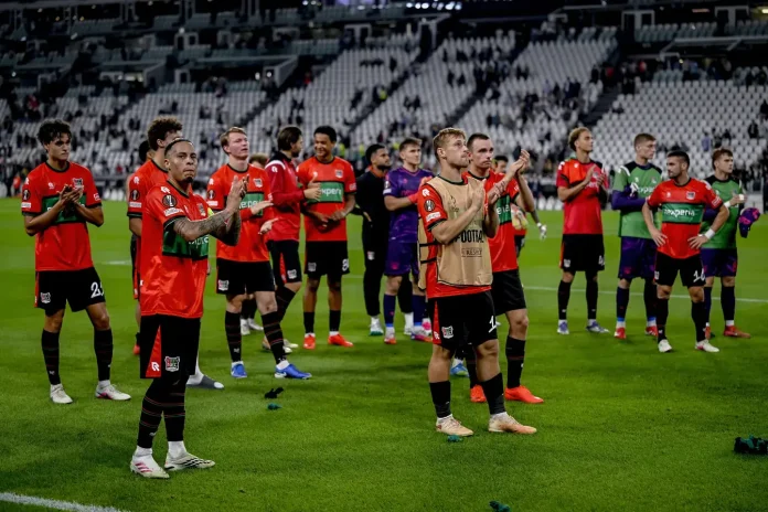 Soccer players in red and black uniforms stand on a field applauding, with stadium seats in the background.