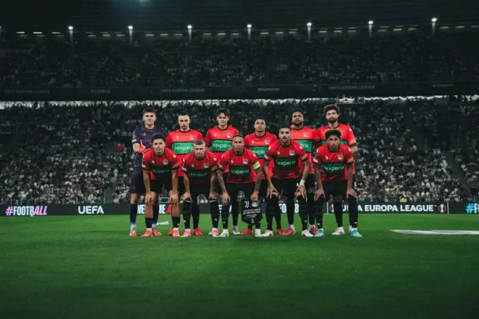 Ten soccer players in red and black jerseys pose for a team photo on a stadium pitch with a packed crowd behind them.