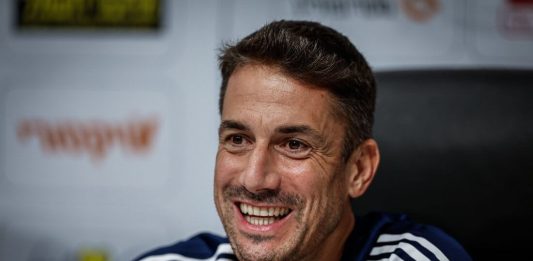 Левски търси голмайстор от висока класа Smiling man with short dark hair and stubble wearing a navy athletic shirt with white stripes, seated in a press/interview setting.