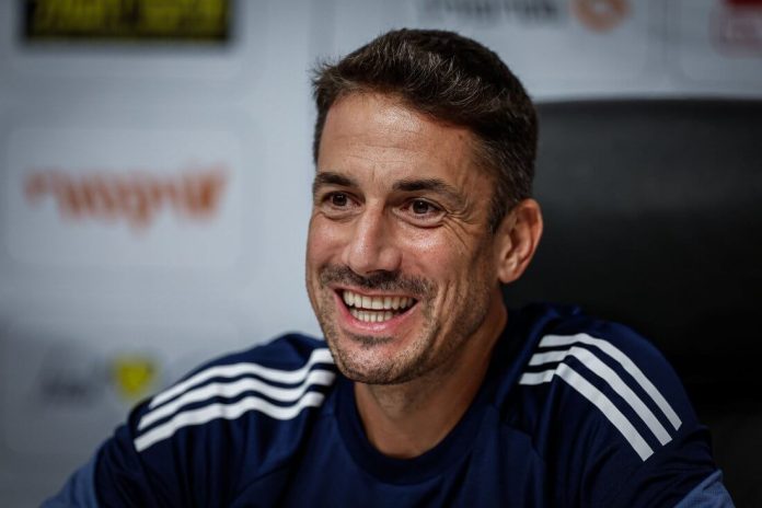 Smiling man with short dark hair and stubble wearing a navy athletic shirt with white stripes, seated in a press/interview setting.