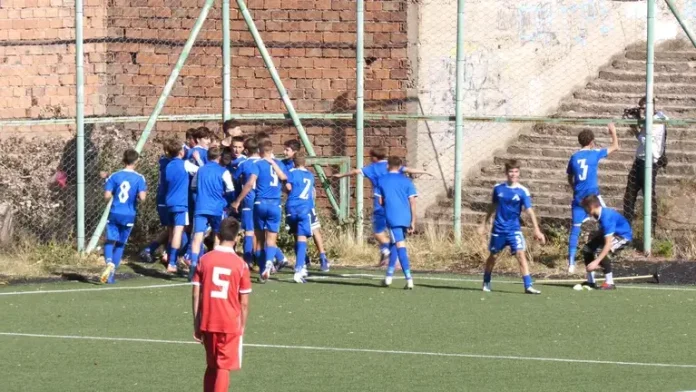 Young soccer players in blue celebrate near the goal on an outdoor field; a red player with number 5 stands in the foreground facing away from the group.