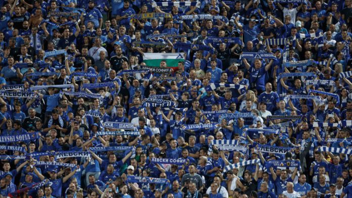 Crowd of blue-clad soccer fans in a stadium, many holding striped scarves overhead.