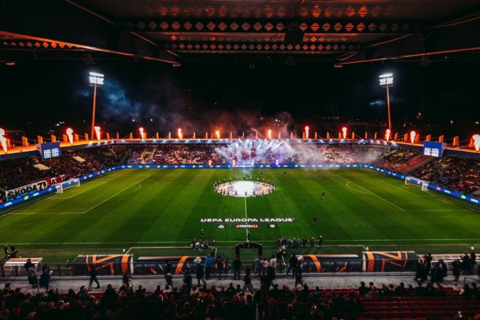 Nighttime UEFA Europa League stadium celebration: circular center-circle display with players around the emblem, flames and smoke in stands, full stands.
