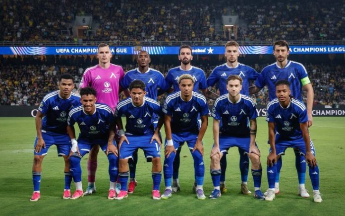 Soccer team in blue kits posing for a pre-match team photo on the field with a UEFA Champions League banner in the background