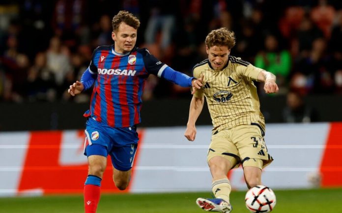 Two soccer players chase the ball during a match; one wears a blue-red striped jersey, the other a beige kit, on the pitch.
