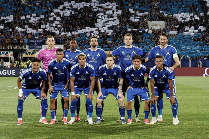 Eleven soccer players in blue uniforms pose for a team photo on the field before a match, with fans in the stands behind them.