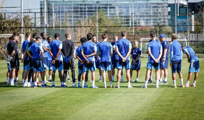 Soccer players in blue training kits stand in a circle on a green grass field during practice with a goal in the background.
