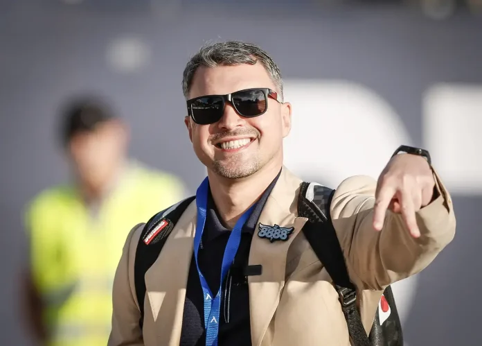 Smiling man in sunglasses and a tan jacket with a blue lanyard, pointing at the camera.