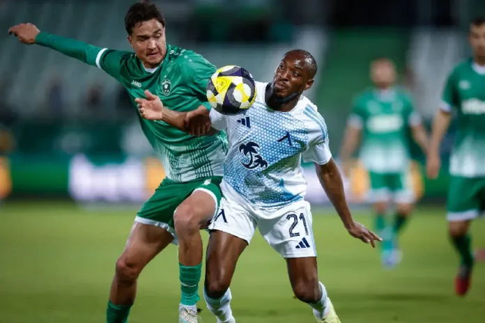Two soccer players vie for the ball during a match, one in green kit and the other in white with blue accents, both leaning in as they challenge for possession.