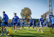 Левски затваря тренировките преди гостуването на Арда Group of soccer players in blue training kits running through a drill on a sunny grass field with yellow cones and a tall stadium structure in the background.