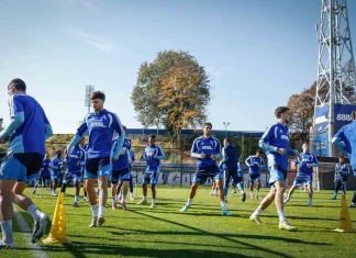 Левски затваря тренировките преди гостуването на Арда Group of soccer players in blue training kits running through a drill on a sunny grass field with yellow cones and a tall stadium structure in the background.