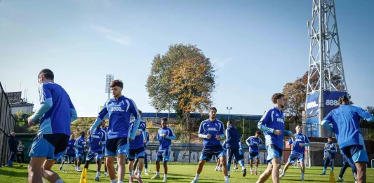 Левски затваря тренировките преди гостуването на Арда Group of soccer players in blue training kits running through a drill on a sunny grass field with yellow cones and a tall stadium structure in the background.
