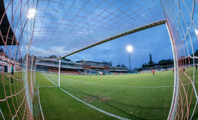 View from inside a soccer goal looking out over a green field with players and a spectator stand in the distance under bright stadium lights at dusk.