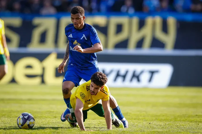 Soccer clash: a blue-team player challenges a yellow-team defender for the ball on a sunlit field, both near the ball.