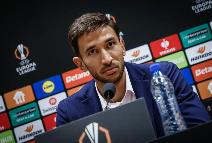 Man in a navy suit sits at a press conference table with a microphone in front of a sponsor-filled backdrop (EUROPA LEAGUE logos visible).