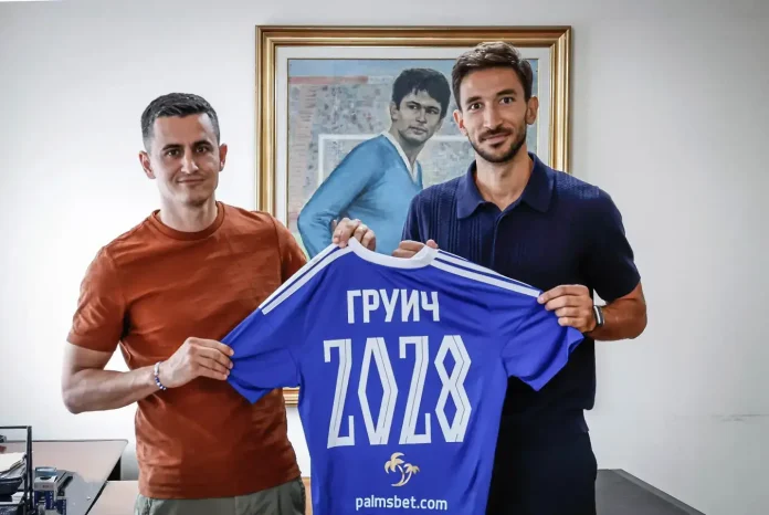 Two men stand side by side holding a blue sports jersey with the number 2028 and Cyrillic text, smiling at the camera in a room with a framed painting behind them.