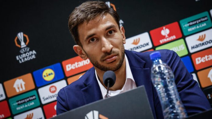 Man in a navy suit speaks into a microphone at a press conference, with a backdrop of sponsor logos behind him. It also shows a water bottle on the table.
