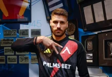 Матей Маркович: история, кариера и отличия Athlete in a Palms Bet jersey poses with his hand extended, in front of a wall of photos and trophies.