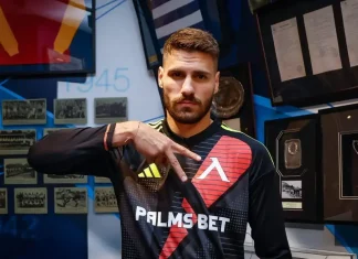 Матей Маркович: история, кариера и отличия Athlete in a Palms Bet jersey poses with his hand extended, in front of a wall of photos and trophies.