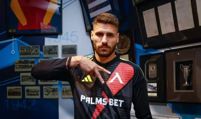 Athlete in a Palms Bet jersey poses with his hand extended, in front of a wall of photos and trophies.
