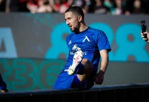 Мазир Сула: история, числа и успехи Male soccer player in a blue kit stepping onto a barrier during a match, crowd blurred in the background.