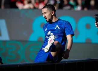 Мазир Сула: история, числа и успехи Male soccer player in a blue kit stepping onto a barrier during a match, crowd blurred in the background.