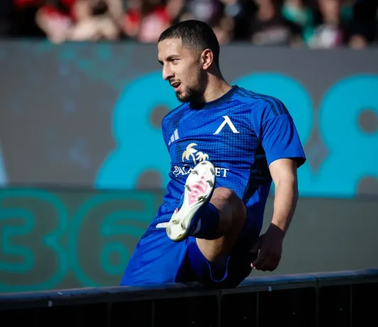 Мазир Сула: история, числа и успехи Male soccer player in a blue kit stepping onto a barrier during a match, crowd blurred in the background.