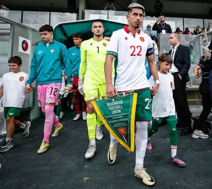 Football players walk through a tunnel, holding a Bulgarian Football Union pennant, as teammates and kids accompany them before a match.