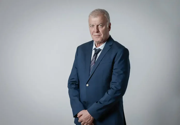 Older man in a navy suit and striped tie posing against a light gray studio backdrop, hands clasped in front.