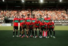 НЕК има успешна история срещу балкански отбори Football team in red and black kit posing for a group photo on the pitch with fans in stands behind them, before a match.