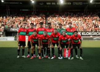 НЕК има успешна история срещу балкански отбори Football team in red and black kit posing for a group photo on the pitch with fans in stands behind them, before a match.