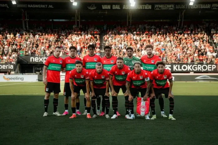Football team in red and black kit posing for a group photo on the pitch with fans in stands behind them, before a match.