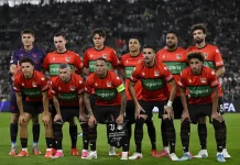 НЕК вдига каре за мача с Левски от Лига Европа NEC Nijmegen football players in red and green kits pose for a group photo on the pitch with a stadium crowd behind them, including a purple-clad goalkeeper on the left.